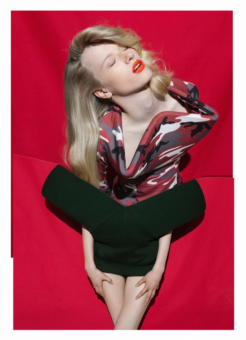 Fashion-forward image of a fair-skinned woman in camouflage top, dark green skirt against a vibrant red backdrop, editorial style.