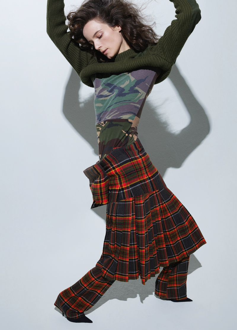 A woman poses indoors wearing a cropped green sweater, camouflage top, and plaid pants with black heeled shoes against a grey background.