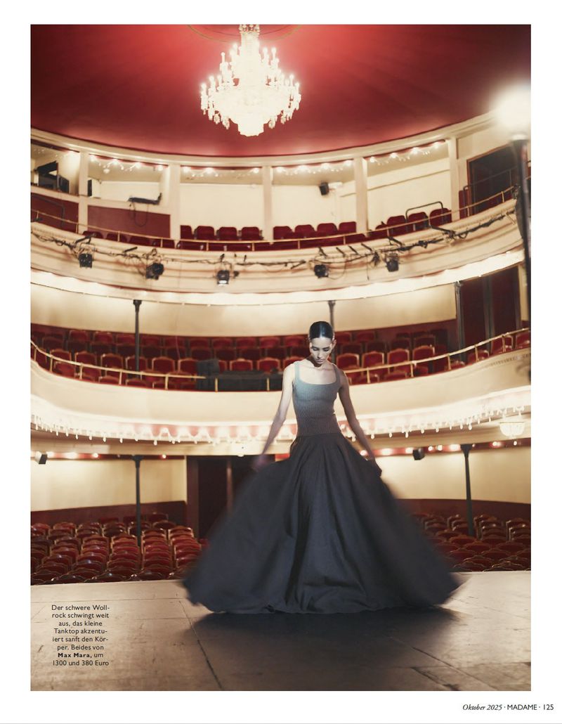 Fashion editorial shot of a woman wearing a grey bodice and flowing black gown, posing on a stage in an elaborate vintage theatre.
