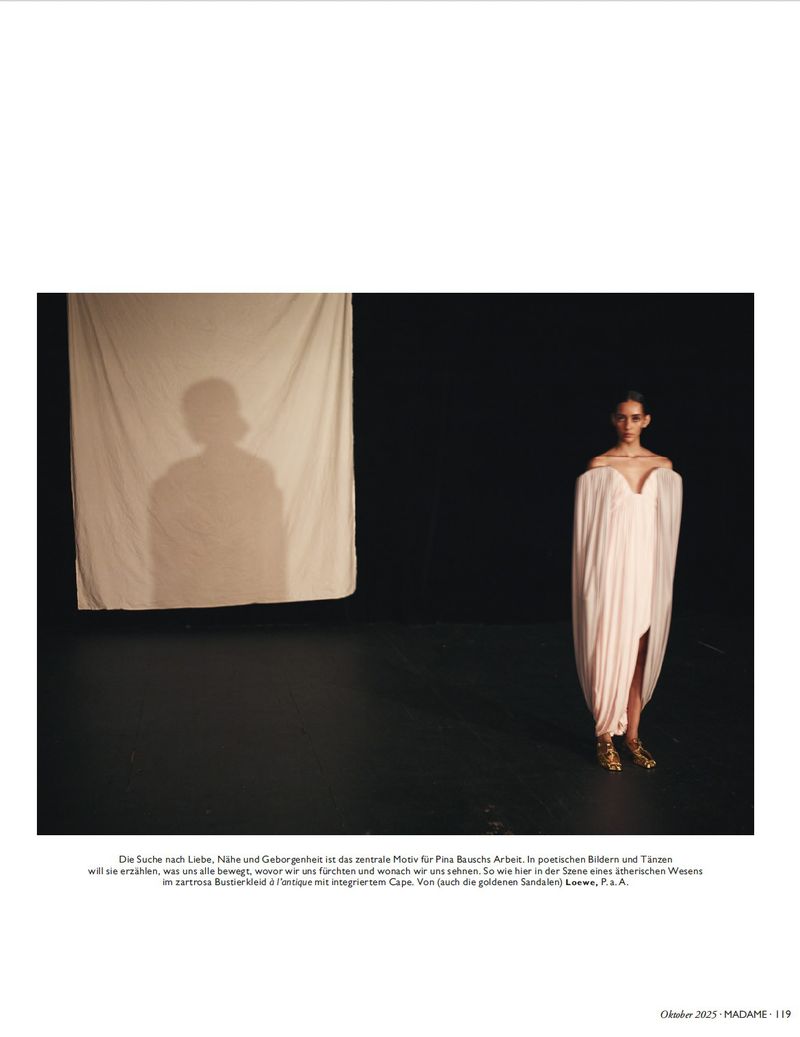 Woman in draped dress poses in dark photo studio with shadow backdrop, showcasing ethereal fashion and studio photography techniques.