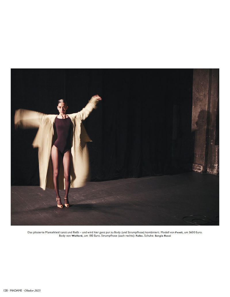 A woman in a leotard and coat poses in a dimly lit studio with her arms outstretched, casting blurred shadows on the dark backdrop.