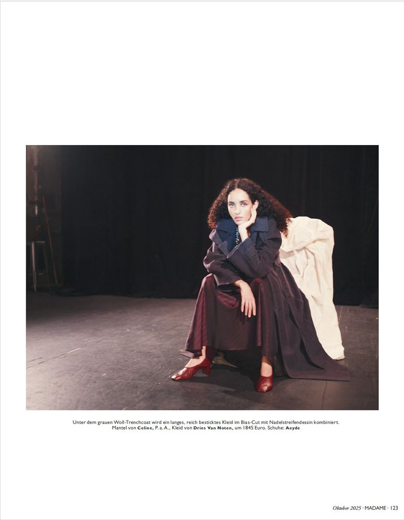 A woman in a long dress and coat sits in a photo studio against a dark backdrop, with her hand resting on her face in a thoughtful pose.