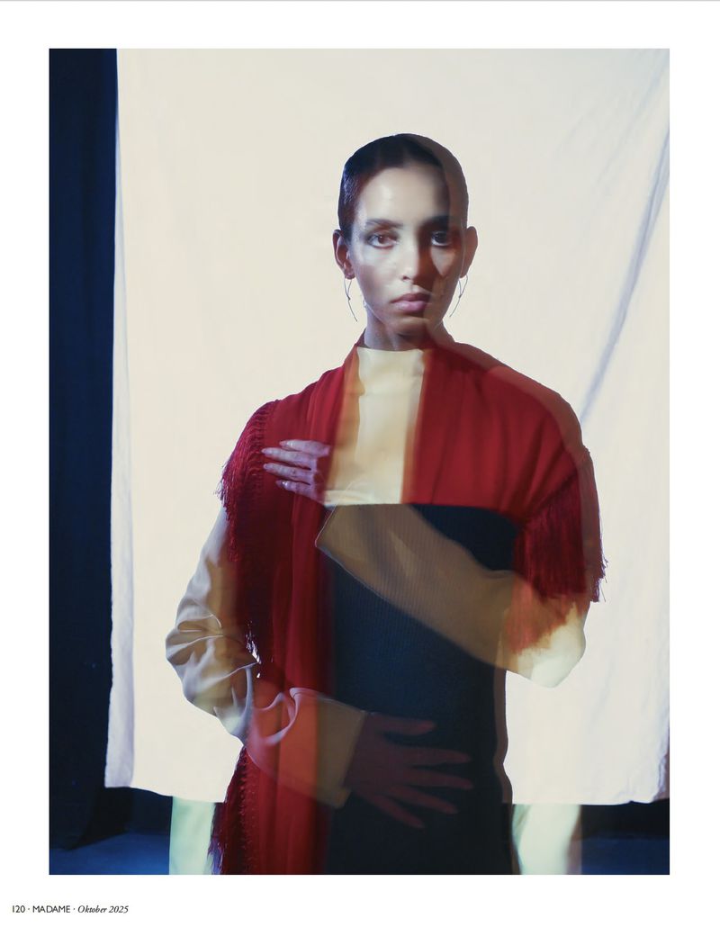 Fashion editorial portrait of a young adult woman with red fringed shawl in front of a light studio background with soft, diffused lighting.