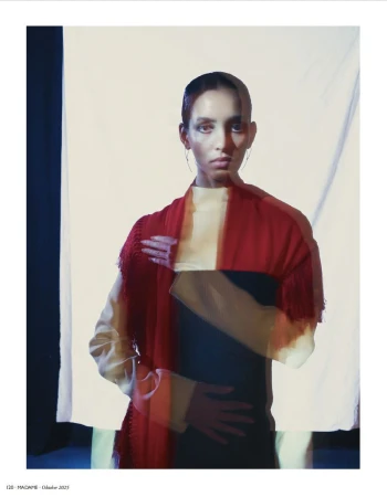 Fashion editorial portrait of a young adult woman with red fringed shawl in front of a light studio background with soft, diffused lighting.