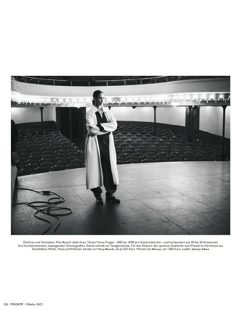 Monochrome shot of a woman in a coat on a theatre stage with rows of empty seats, bathed in soft, natural light.