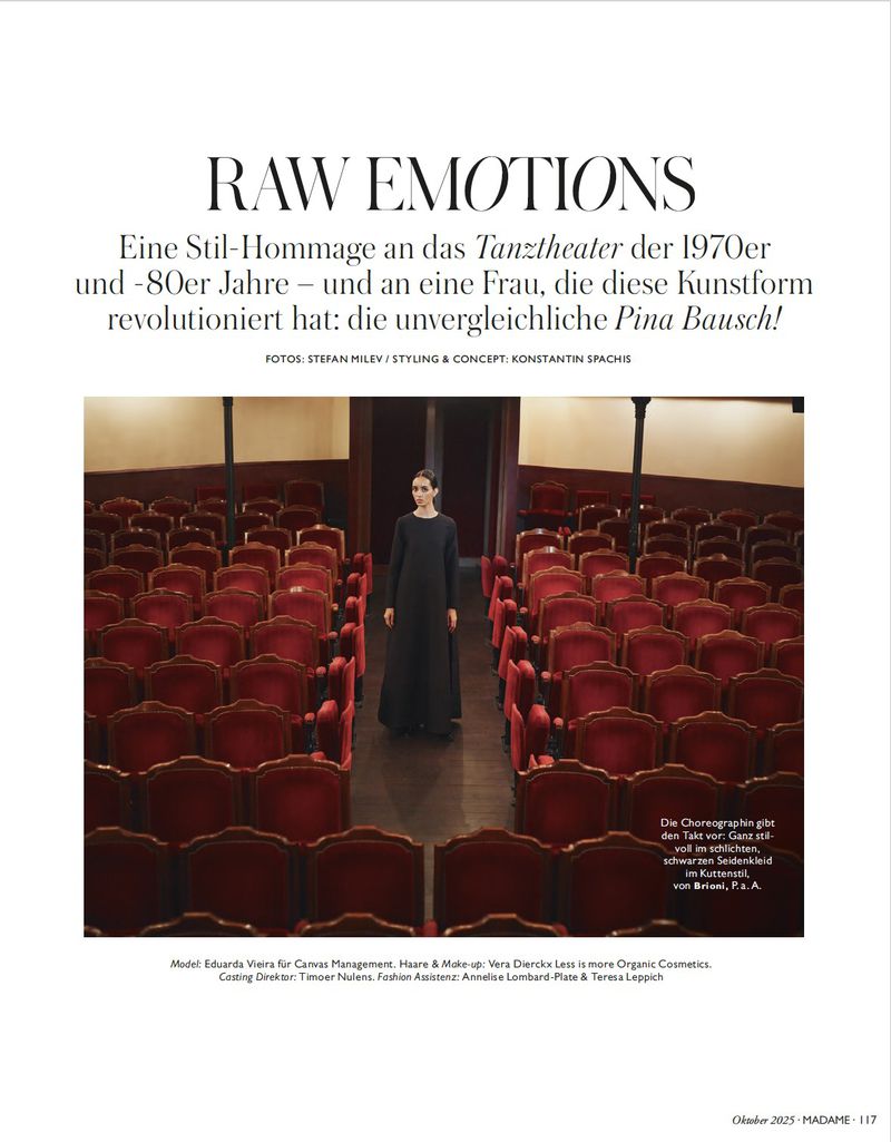 Fashion editorial shot: a single female figure in a black dress stands at the end of an aisle of red theatre seats in an editorial scene.