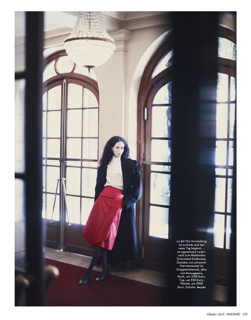 Fashion editorial shot of a woman in a white turtleneck, red skirt, and black coat posing indoors with arched windows and chandelier.