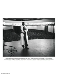 Model standing with arms crossed on a theater stage wearing a light coat and dark trousers in a black and white editorial photograph.