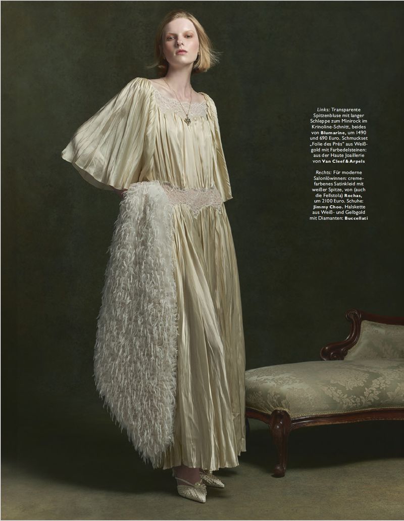 Woman in flowing gown with feather boa poses against dark green backdrop near antique chair in a fashion editorial style portrait.