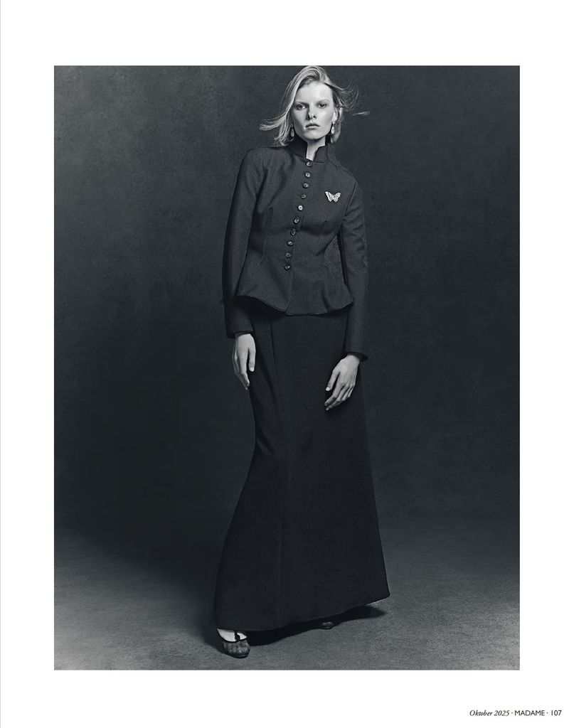 Monochrome portrait of a blond woman in a dark jacket and long skirt against a dark backdrop in a studio setting.