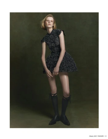 A fashionable woman in a black dress and knee socks poses in a studio. The backdrop is a solid, muted green tone.