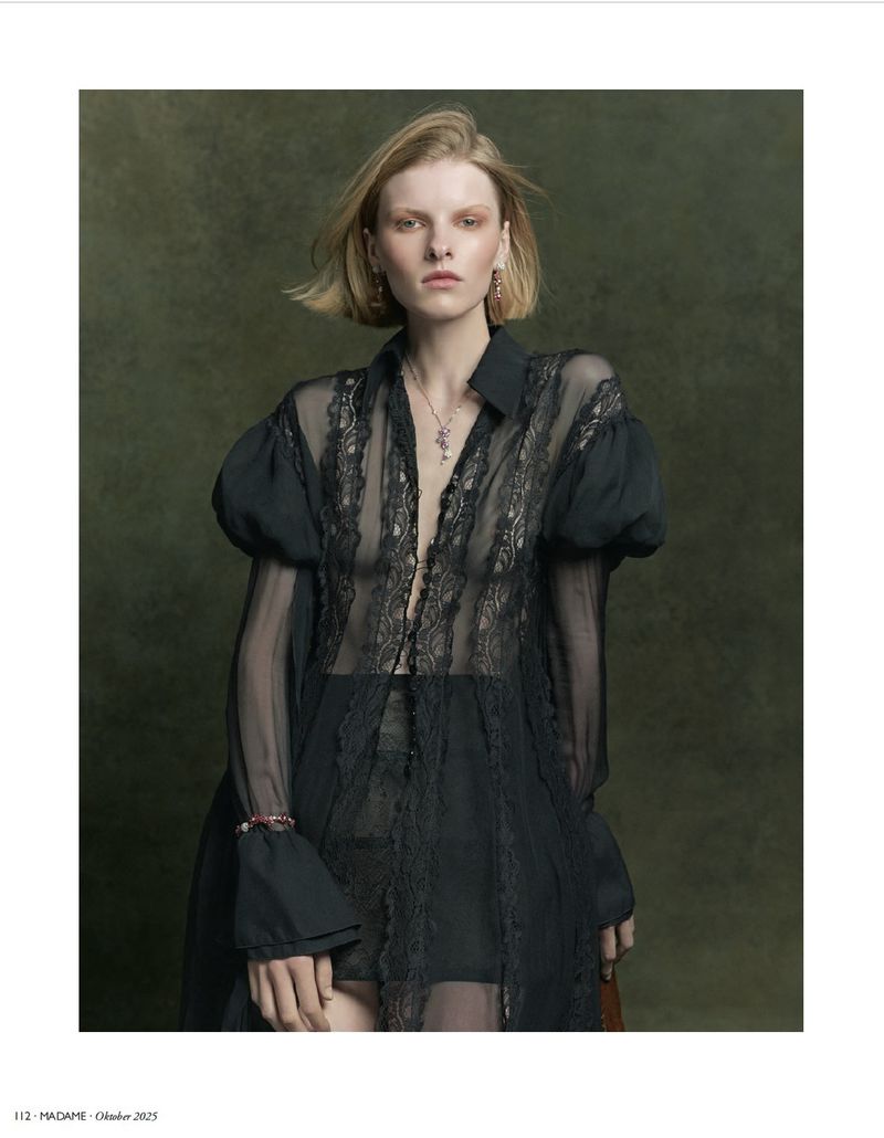 Blonde woman in a black lace shirt with voluminous sleeves poses indoors against a textured green backdrop in a medium shot.