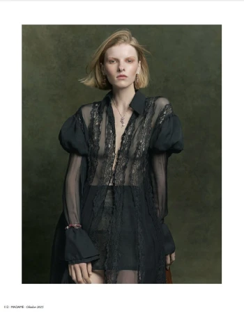 Blonde woman in a black lace shirt with voluminous sleeves poses indoors against a textured green backdrop in a medium shot.