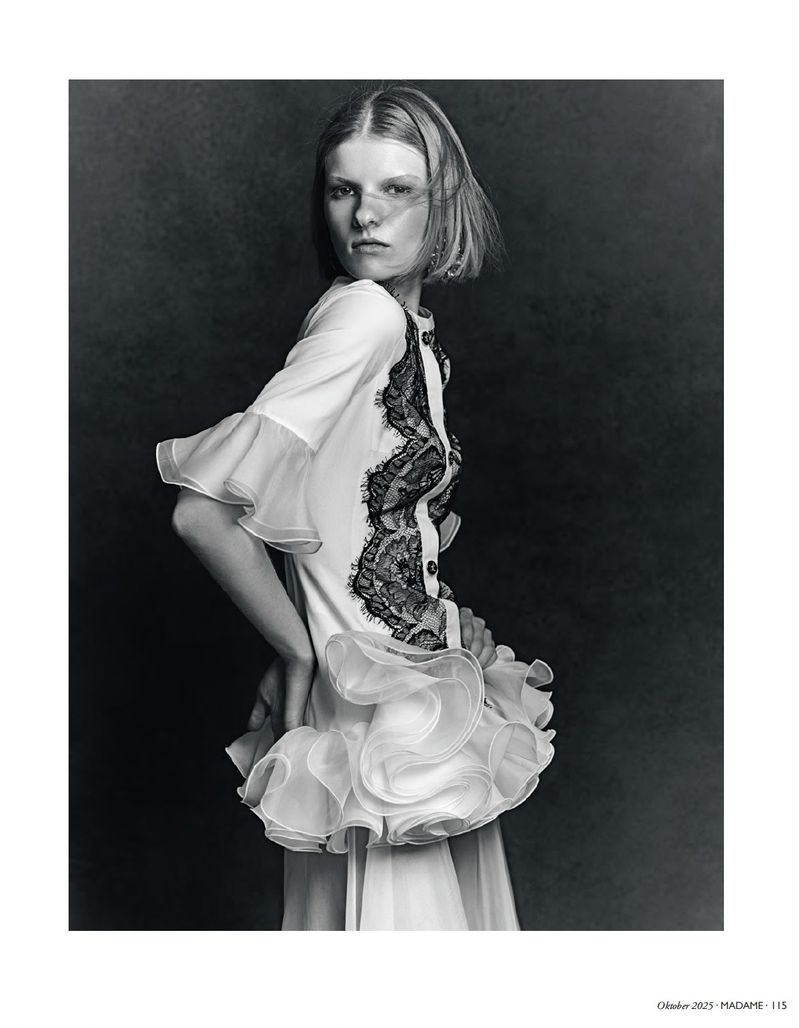 Black and white portrait of a young woman in white ruffled dress with lace against dark background in a studio setting, hands on hip.