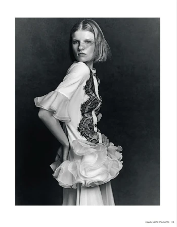 Black and white portrait of a young woman in white ruffled dress with lace against dark background in a studio setting, hands on hip.