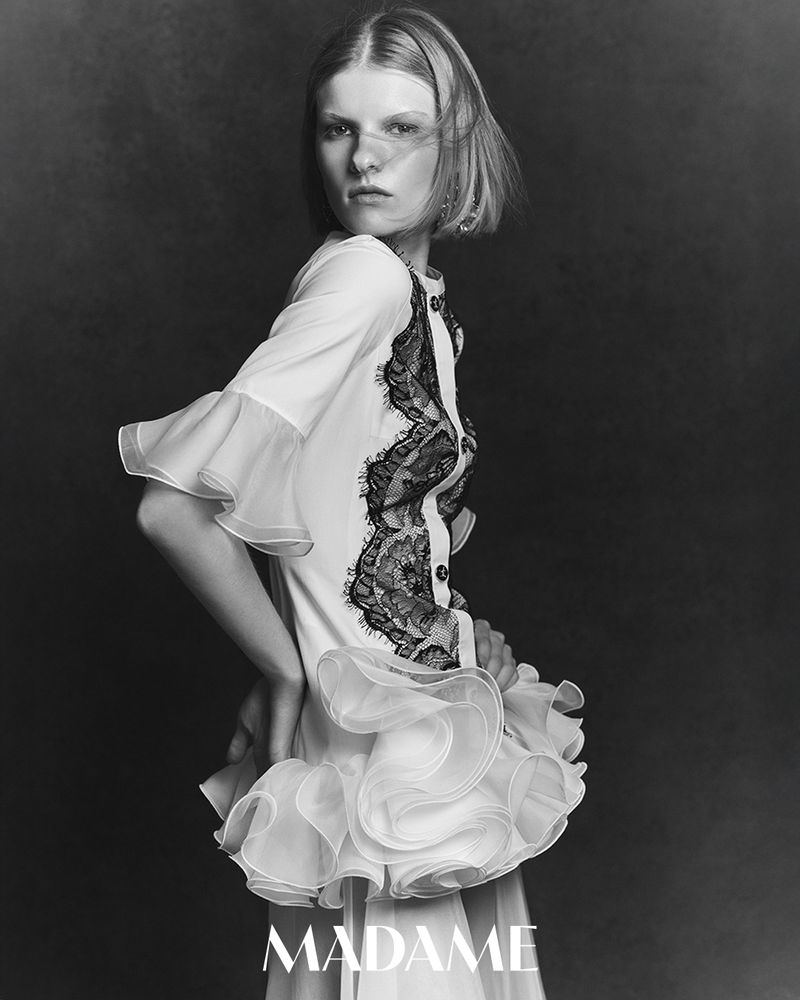Black-and-white portrait of a slender woman with short hair wearing a white dress with lace and ruffles, against a dark grey backdrop.