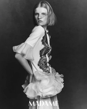 Black-and-white portrait of a slender woman with short hair wearing a white dress with lace and ruffles, against a dark grey backdrop.