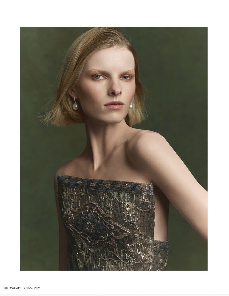 A fashionable portrait of a fair-skinned model wearing a beaded strapless dress against a neutral green background, showcasing beauty and style.