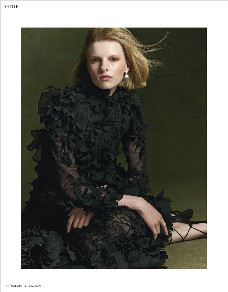 A fair-skinned woman with blonde bobbed hair poses in a black lace dress with rosette details in a studio with a dark green background.