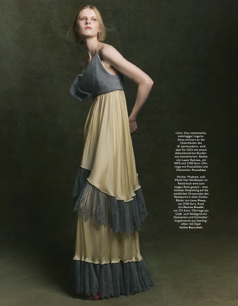 A fair-skinned woman in a tiered, flowing maxi dress stands indoors against a mottled green backdrop in a fashion editorial shot.
