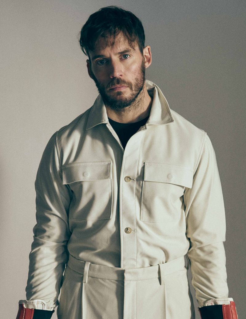 Portrait of a handsome adult male with a beard, wearing a cream-colored jacket and pants, shot in a photo studio with neutral lighting.