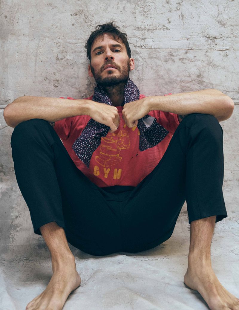 An adult man with a beard, wearing a red t-shirt and black pants, is sitting on a textured surface against a grey textured wall.