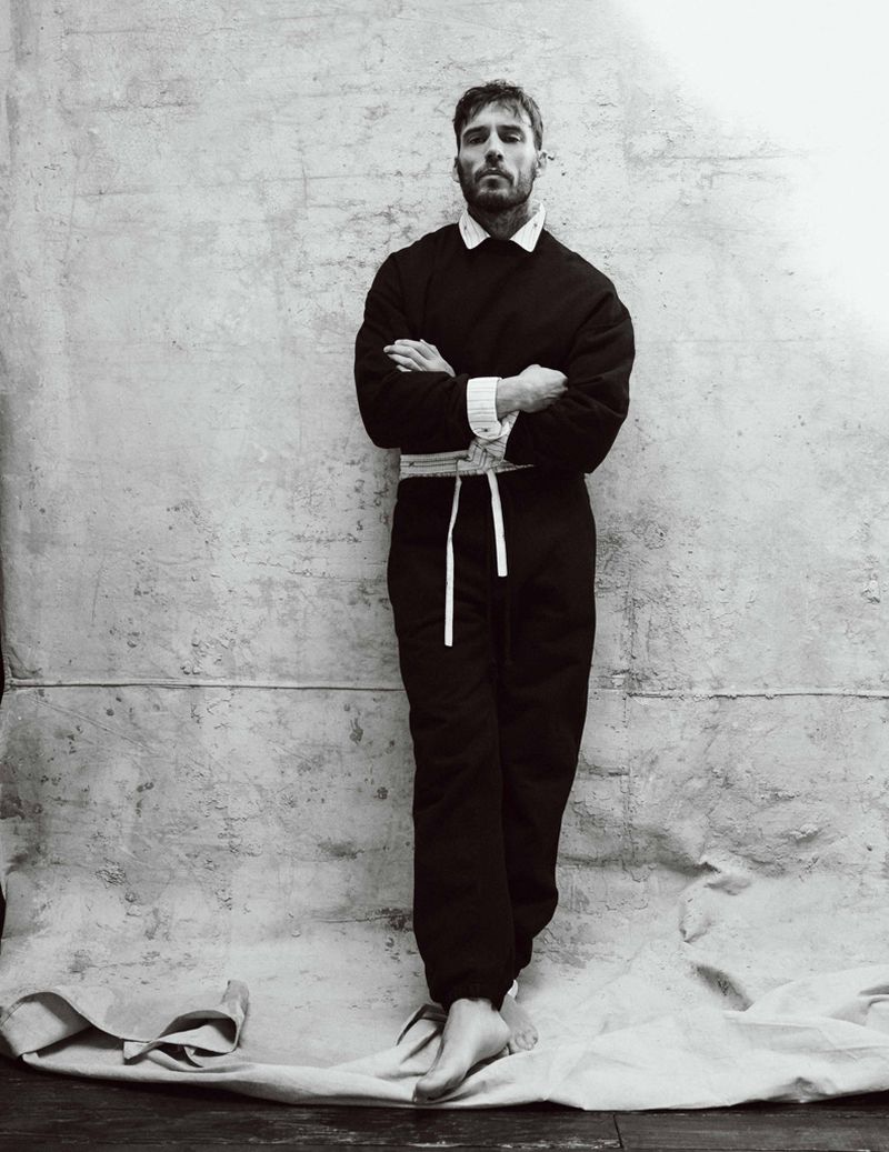 Sam Claflin standing barefoot with arms crossed in a dark belted jumpsuit against a textured canvas backdrop.