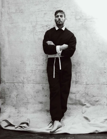 Sam Claflin standing barefoot with arms crossed in a dark belted jumpsuit against a textured canvas backdrop.