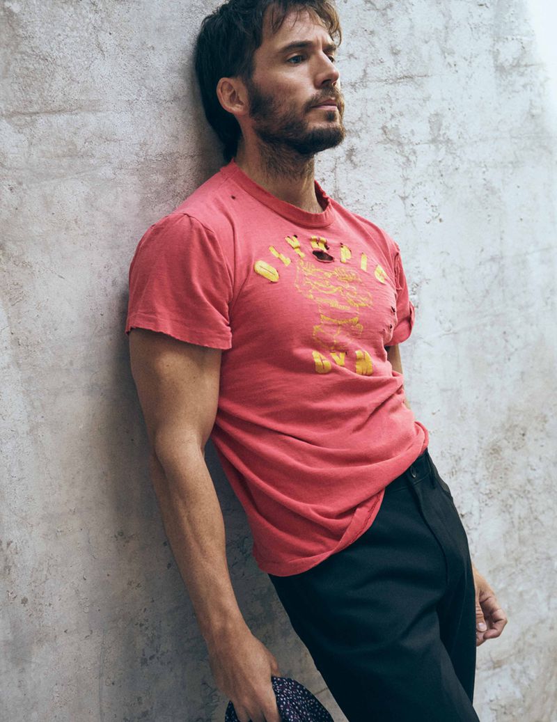 Portrait of a fair-skinned male model with a beard wearing a pink t-shirt and black pants, leaning against a concrete wall in a studio.