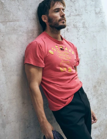 Portrait of a fair-skinned male model with a beard wearing a pink t-shirt and black pants, leaning against a concrete wall in a studio.
