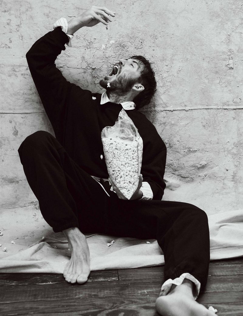 Black and white portrait of a bearded man sitting while holding a bag of popcorn and throwing some into his open mouth.
