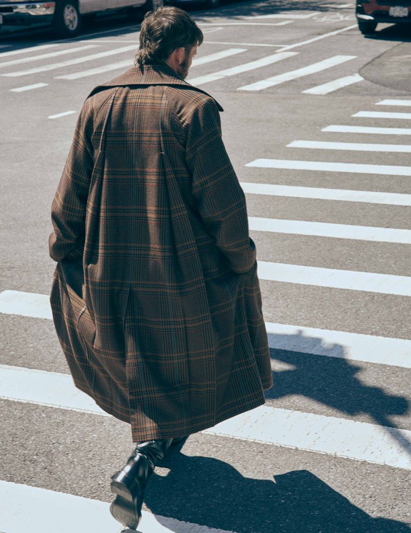 Actor Sam Claflin seen from behind walking across a city crosswalk wearing a long brown plaid overcoat and black boots.