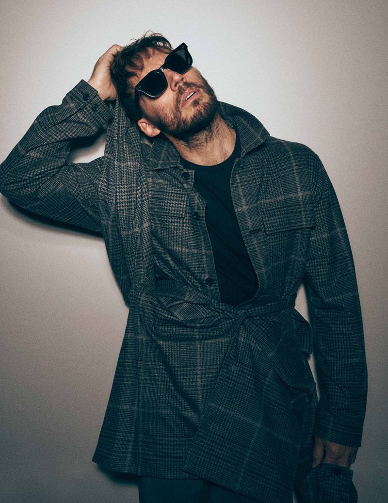 Stylish adult male in sunglasses poses indoors, wearing a grey plaid jacket over a black shirt, hand on head, looking up.