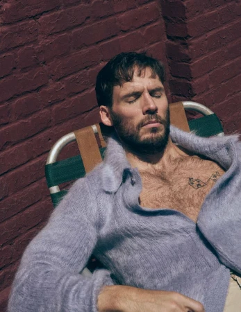 Sam Claflin reclining in a lawn chair with eyes closed wearing an unbuttoned lavender mohair cardigan against a brick wall.