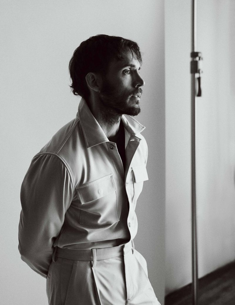 Sam Claflin in a light collared shirt and trousers standing near a studio light stand in a black and white portrait.