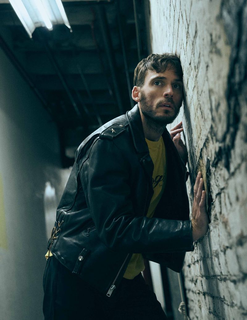 Man in a leather jacket leans against a brick wall in dim light, looking off-camera with a serious expression in an urban environment.
