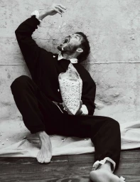 Black and white photo of Sam Claflin sitting barefoot on the floor, catching falling popcorn in his mouth from a clear bag.