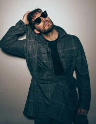 Sam Claflin wearing dark sunglasses and a plaid shirt, posing against a light grey studio background for Flaunt.