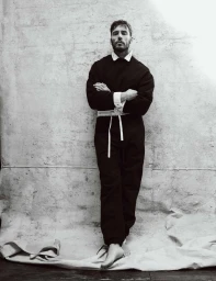 Sam Claflin standing barefoot with arms crossed in a dark belted jumpsuit against a textured canvas backdrop.