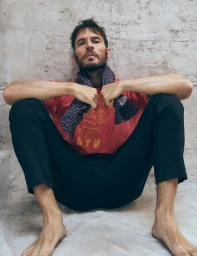 Sam Claflin sitting against a textured concrete wall wearing a red graphic t-shirt, dark trousers, and a patterned scarf.