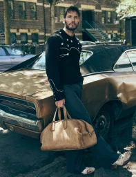 Actor Sam Claflin wearing a black embroidered sweater and holding a tan suede duffle bag while leaning against a vintage car.