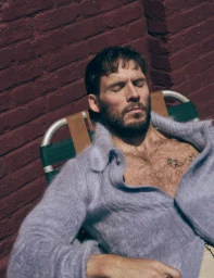 Sam Claflin reclining in a lawn chair with eyes closed wearing an unbuttoned lavender mohair cardigan against a brick wall.