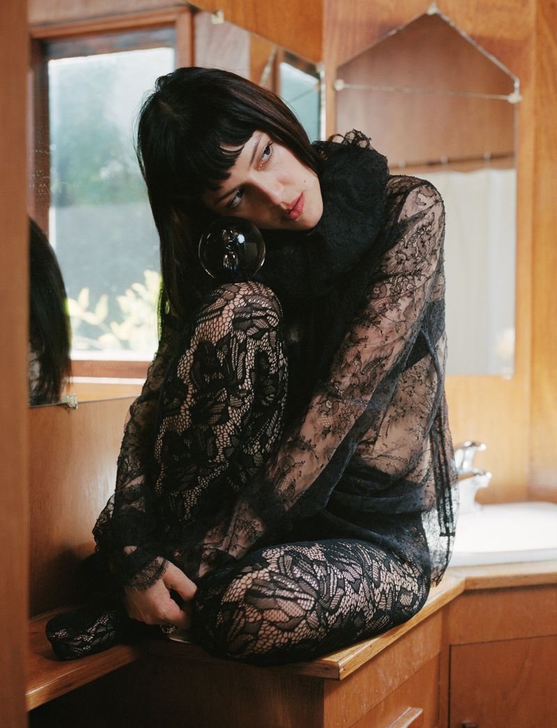Adult female in black lace top and pants sits on a counter in an indoor environment with warm lighting and a mirror.
