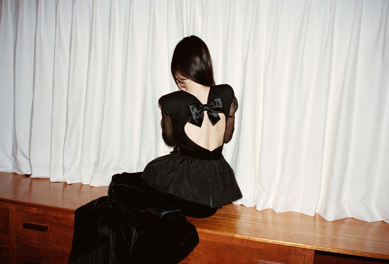 Back view of an adult female in black fashion with a bow, seated indoors against a white curtain backdrop.