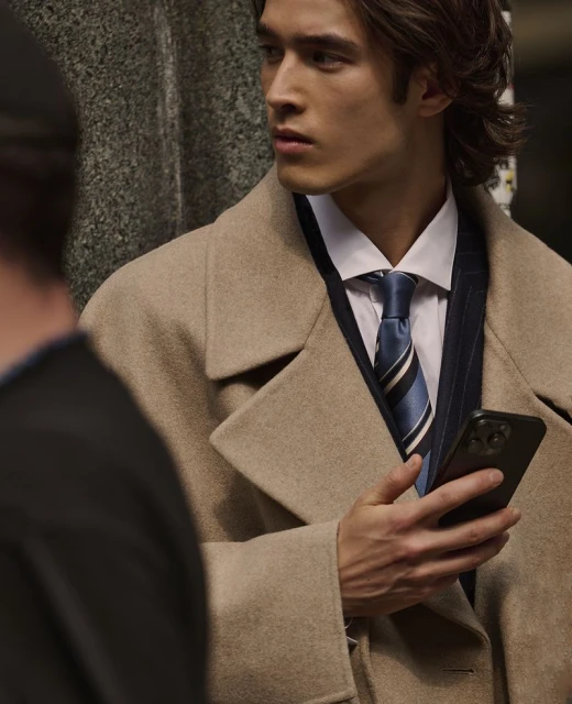 Asian male model in a beige coat holding a smartphone, looking away in an outdoor setting with a neutral color palette.
