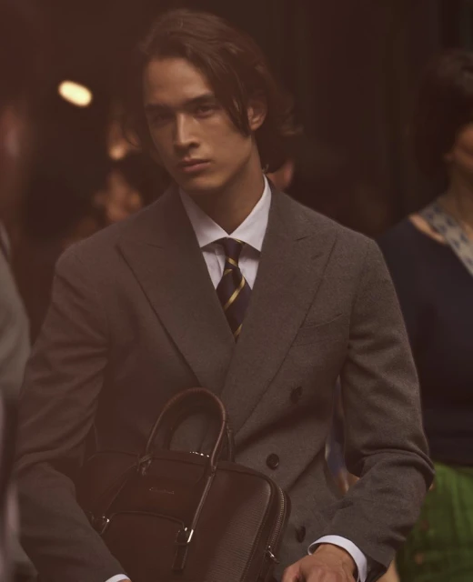 Portrait of a stylish adult male wearing a suit and carrying a briefcase, posed against a blurred urban backdrop with warm lighting.