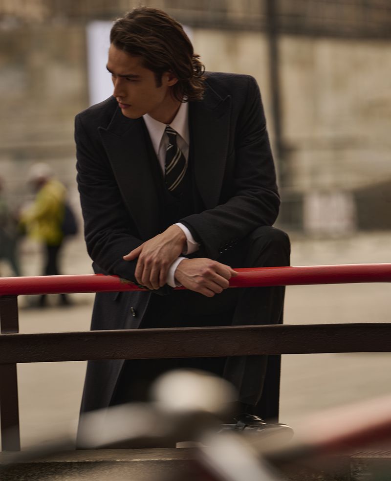Man in dark coat and suit leans against red railing outdoors, looking to the side in a medium shot with soft-focus background.