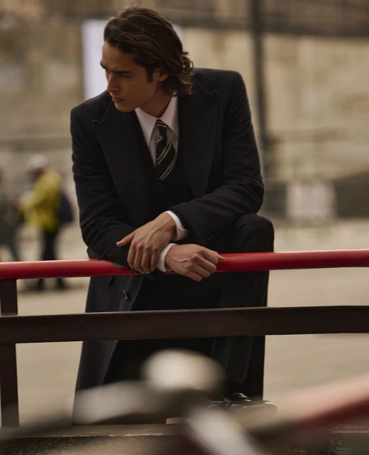 Man in dark coat and suit leans against red railing outdoors, looking to the side in a medium shot with soft-focus background.