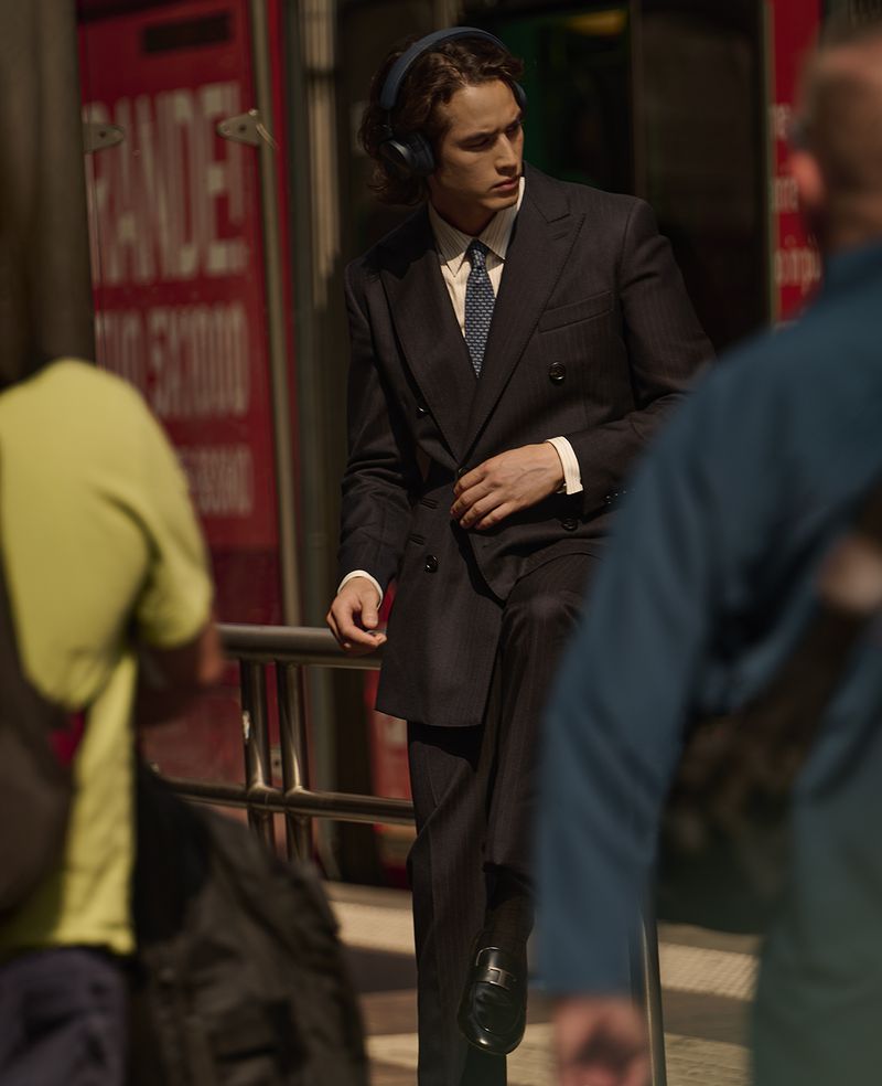 Man in suit wearing headphones leans on a railing in a city setting, featuring blurred pedestrians and urban background, with dark, muted tones.