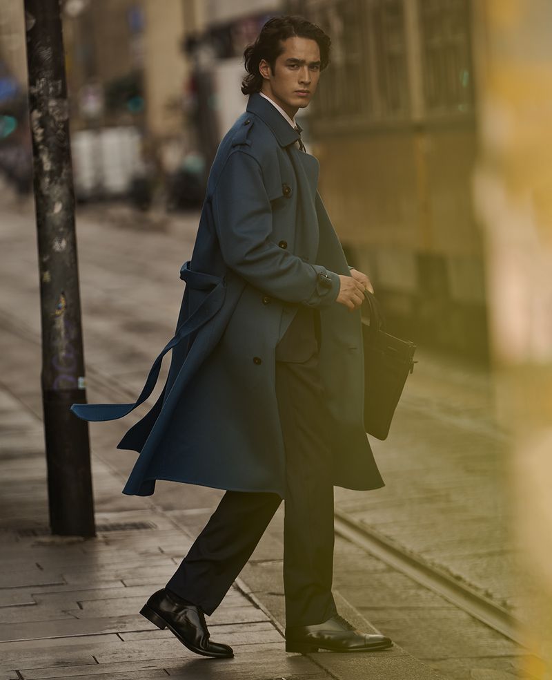 A brunette man in a teal trench coat walks across a city street, carrying a black bag, with blurred buildings in the background.